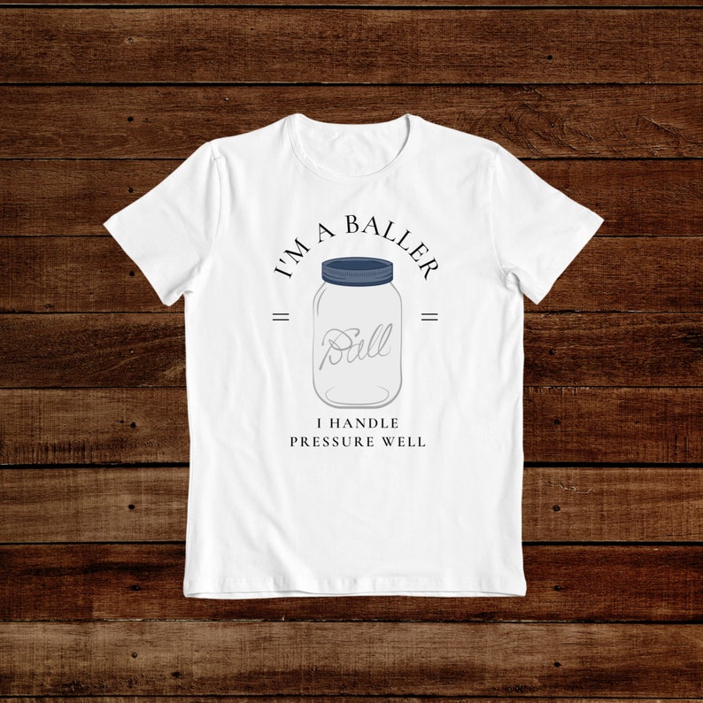 Puede incluir: Camiseta blanca con un gr&aacute;fico de un frasco de vidrio con la palabra "Ball" dentro. El texto "I'm a Baller" est&aacute; por encima del frasco y "I handle pressure well" est&aacute; por debajo del frasco.
