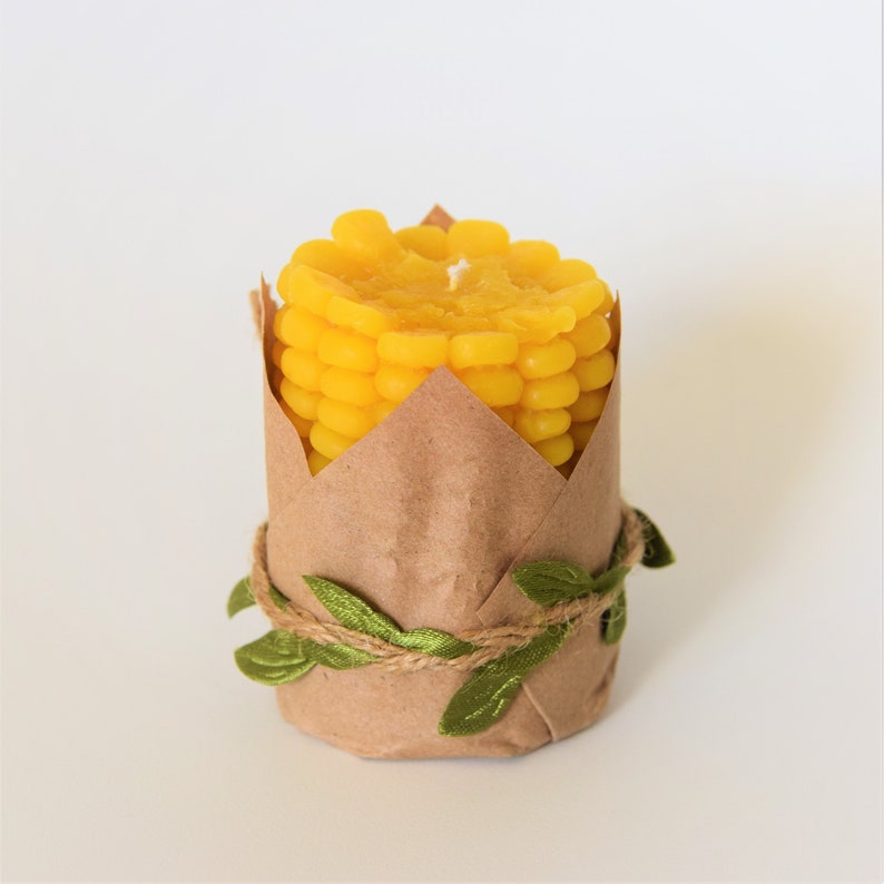 May include: A yellow corn on the cob candle wrapped in brown paper with green leaves and twine.