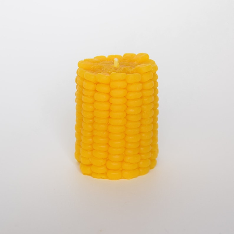 May include: A yellow candle shaped like an ear of corn. The candle has a white wick in the center.