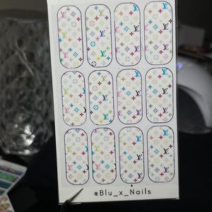 May include: A rectangular sheet of twelve nail wraps featuring a repeating pattern of multicoloured LV-style logos on a white background. The sheet includes the text @Blu_x_Nails printed in black at the bottom centre of the clear plastic packaging.