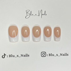 May include: Five nude-colored press-on nails with a white french tip design. The nails are arranged in a row from left to right.
