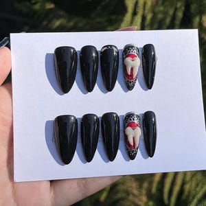 Könnte beinhalten: Satz mit zehn schwarzen, aufklebbaren Nägeln mit einem roten und weißen Zahndesign. Die Nägel sind in zwei Reihen zu fünf auf weißem Hintergrund angeordnet.