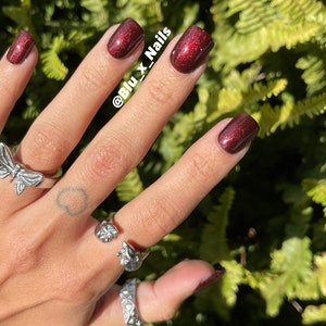Könnte beinhalten: Nahaufnahme einer Hand mit rotem Glitzernagellack. Die Hand trägt mehrere silberne Ringe, darunter einen Schmetterlingsring. Der Hintergrund ist eine grüne, belaubte Pflanze. Der Text "@Blu_x_Nails" ist auf der Hand sichtbar.
