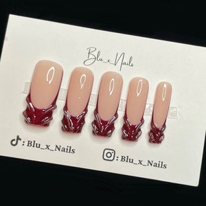 May include: Set of five burgundy and nude false nails with a white design. The nails are displayed on a white card with the text "Blu_x_Nails" and social media handles for TikTok and Instagram.