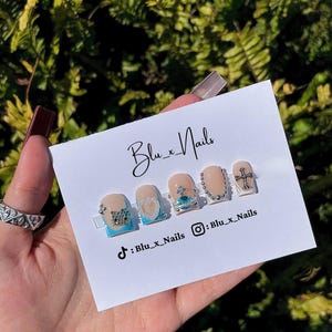 Könnte beinhalten: Ein Set mit fünf kurzen, quadratischen, hellblauen und weißen Nagelspitzen mit silbernen Akzenten. Die Nagelspitzen haben verschiedene Designs, darunter ein Herz, eine Krone, ein Kreuz und ein Design mit Schmucksteinen. Die Nagelspitzen werden auf einer weißen Karte mit dem Text "Blu_x_Nails" und Social-Media-Handles angezeigt.