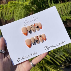 Könnte beinhalten: Set mit künstlichen Nägeln mit Halloween-Motiv. Die Nägel sind nude mit schwarzen und weißen Designs von Katzen, Monden und Sternen. Die Spitzen der Nägel sind orange lackiert.