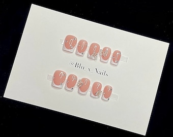 May include: Set of ten nude-colored press-on nails with white tips. Each nail has a silver butterfly design and a single rhinestone. The nails are arranged in two rows on a white background. The text "@Blu_x_Nails" is written below the nails.