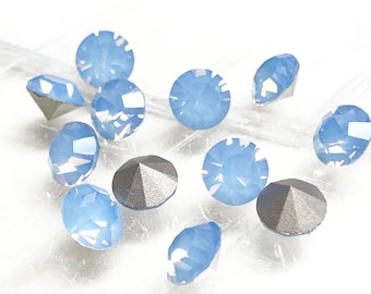 May include: A collection of light blue, faceted gemstones with a round shape. The gems have a shimmering, opalescent quality and are scattered across a white surface. Some gems have a dark gray, metallic backing.