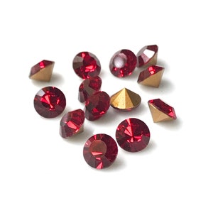 May include: A group of red crystal rhinestones with gold settings. The rhinestones are scattered on a white background.