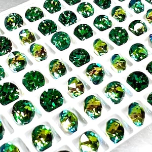 May include: A collection of faceted green gemstones, arranged in a grid within a white tray. The gems vary in shape and have a sparkling, iridescent quality, with hints of yellow and blue within the green. The arrangement suggests a display of jewelry components.