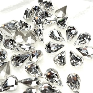 May include: A collection of clear, faceted teardrop-shaped rhinestones scattered on a white surface. The rhinestones have a brilliant, reflective quality, catching the light and creating a sparkling effect. Some are pointed, others are more rounded.