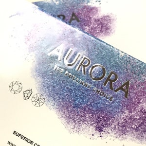 May include: A white box with a silver foil design that says "Aurora The Brilliant Choice" with a watercolor background of purple and blue. The box also has two small diamond illustrations.