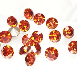 May include: A collection of 15 orange-red rhinestones, each with a faceted design, scattered on a white surface.