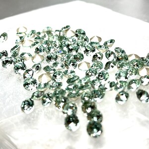 May include: A collection of numerous small, round, faceted gemstones in a pale mint green hue. Each stone features multiple reflective surfaces that catch the light, creating a sparkling effect across the cluster of loose, circular, translucent gems.