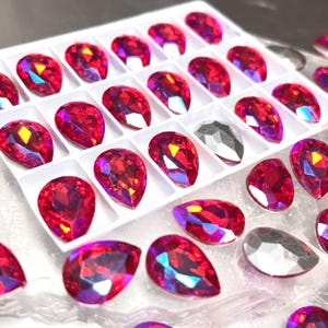 May include: A tray of faceted, teardrop-shaped rhinestones in shades of red and silver. The red stones have a vibrant, iridescent quality, reflecting light with hints of yellow and blue. The silver stones are a neutral accent.
