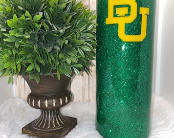 May include: A green glitter tumbler with a yellow "BU" logo. The tumbler is sitting on a white surface next to a potted plant.