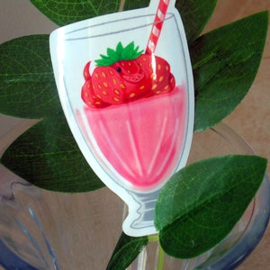 Op de afbeelding: Een sticker met een roze aardbeienmilkshake in een helder glas met een rood-wit gestreept rietje. De aardbei is rood met gele zaden en groene bladeren. De sticker is omgeven door groene bladeren.