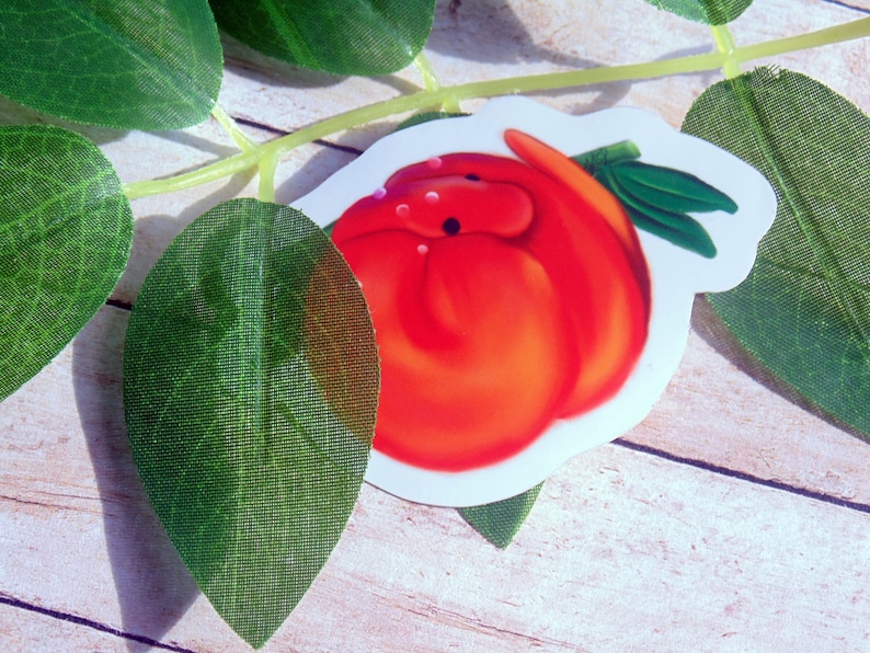 Puede incluir: Una pegatina roja y blanca de un gusano de dibujos animados con una hoja verde en la boca. La pegatina est&aacute; sobre una superficie de madera con hojas verdes.