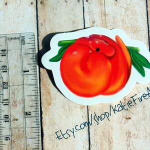 Puede incluir: Una pegatina de melocot&oacute;n naranja brillante con un tallo y hojas verdes. La pegatina est&aacute; sobre un fondo blanco y tiene un ligero brillo. El texto "Etsy.com/shop/KatieFireArt" est&aacute; escrito en negro en la parte inferior de la pegatina.