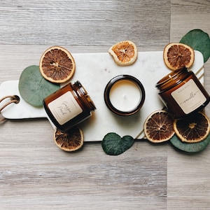 Può includere: Tre barattoli di vetro ambrato per candele con coperchi su una superficie di marmo bianco. Le candele sono etichettate "Vaniglia", "Gardenia Peach" e una è senza etichetta. Fette di arancia essiccate e foglie verdi circondano le candele.
