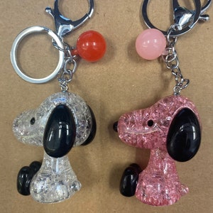 May include: Two keychains featuring a cartoon dog design. One keychain has a clear, sparkly, white dog with black ears and a red bead. The other keychain has a clear, sparkly, pink dog with black ears and a pink bead.