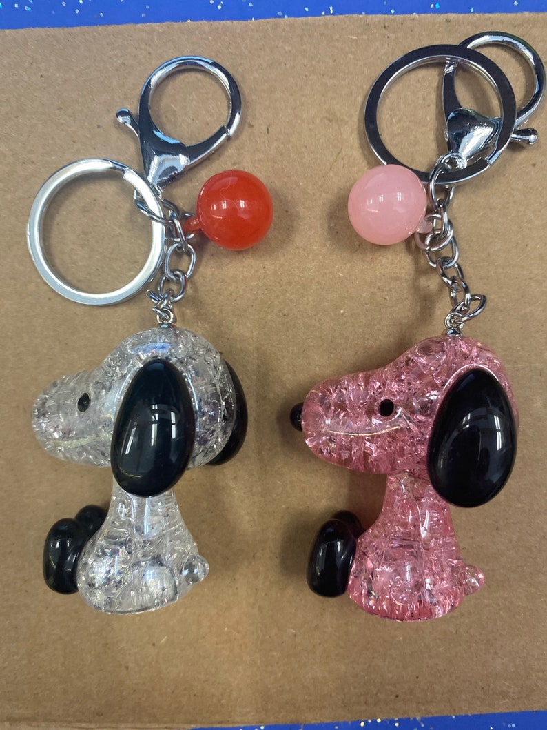 May include: Two keychains featuring a cartoon dog design. One keychain has a clear, sparkly, white dog with black ears and a red bead. The other keychain has a clear, sparkly, pink dog with black ears and a pink bead.