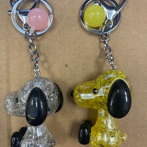 May include: Two keychains featuring a cartoon dog with a sparkly finish. One keychain is silver with a pink charm and the other is yellow with a yellow charm.