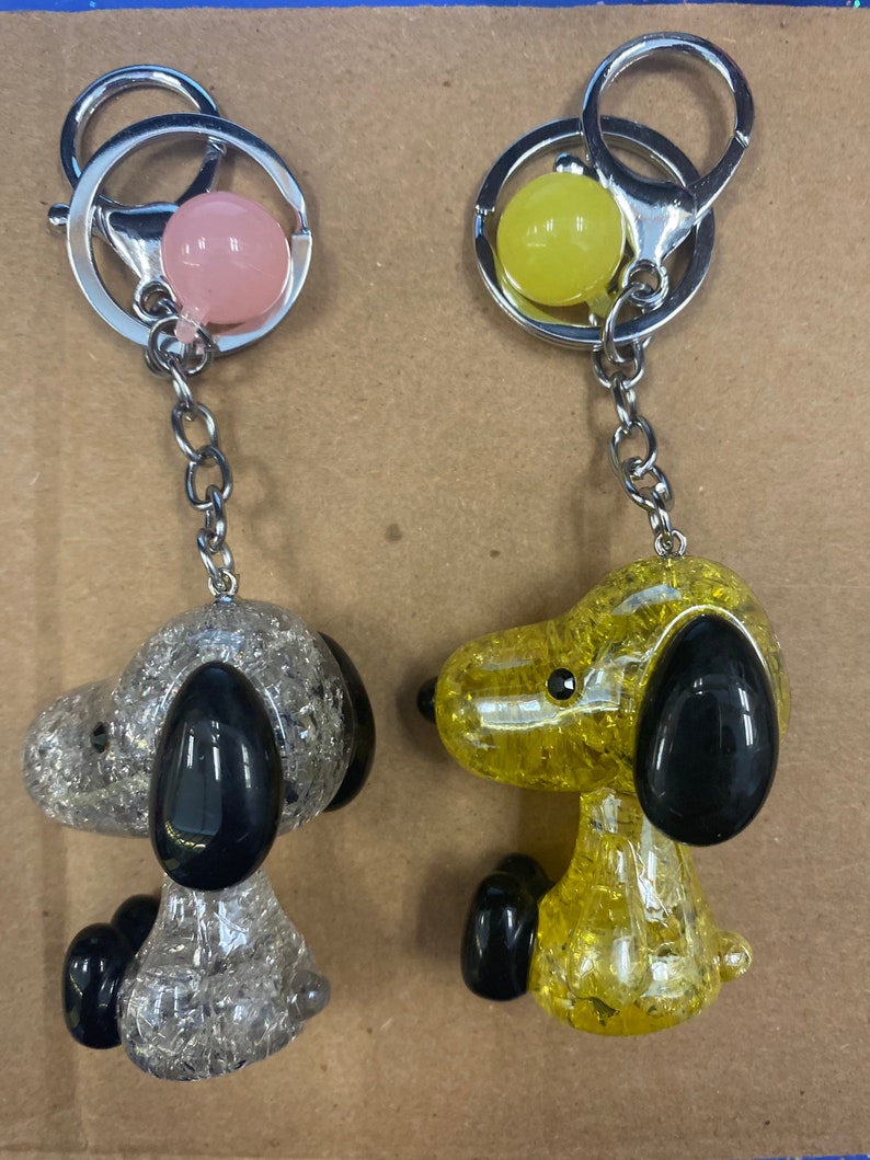 May include: Two keychains featuring a cartoon dog with a sparkly finish. One keychain is silver with a pink charm and the other is yellow with a yellow charm.
