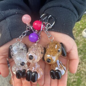 May include: Three glass dog keychains with glitter. The dogs are white, pink, and gold. Each keychain has a black nose and black feet. The keychains are attached to a silver chain with pink, purple, and red beads.