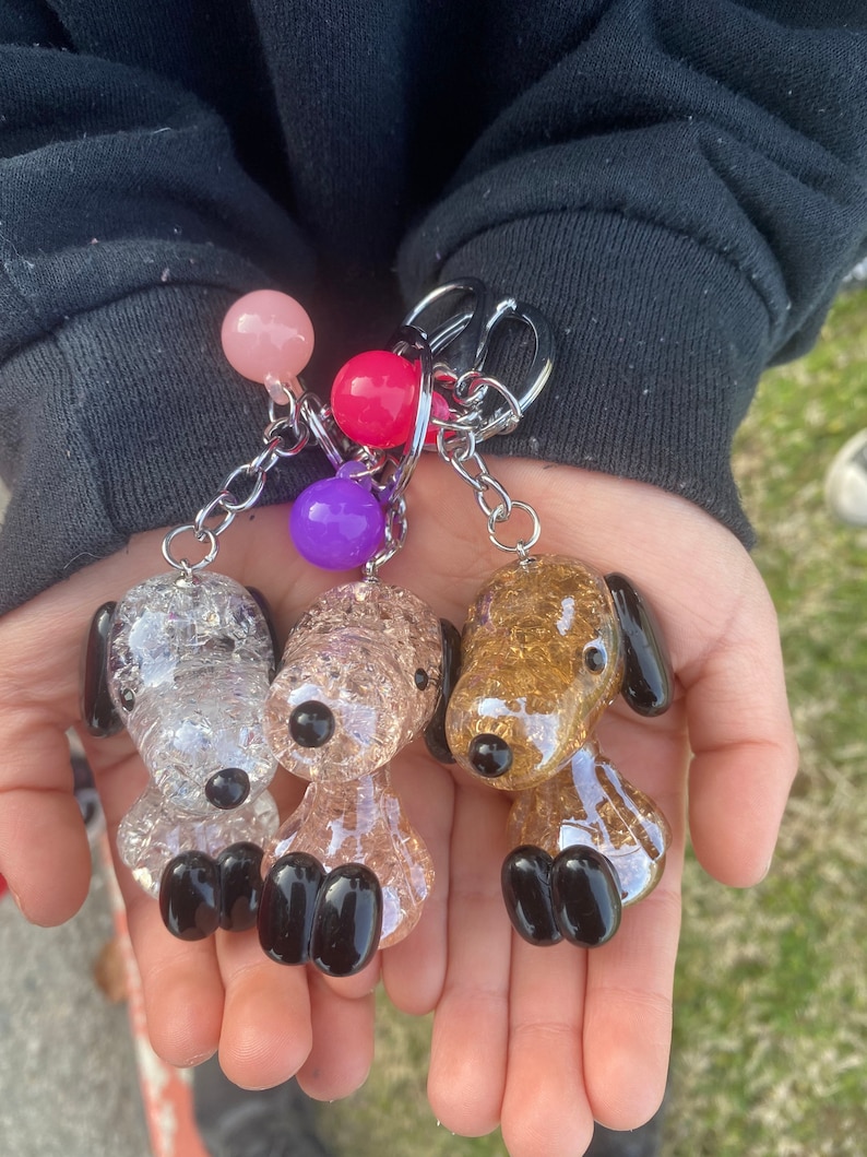 May include: Three glass dog keychains with glitter. The dogs are white, pink, and gold. Each keychain has a black nose and black feet. The keychains are attached to a silver chain with pink, purple, and red beads.