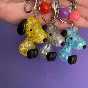 May include: Three keychains featuring a cartoon dog design. The dogs are made of clear glass with glitter embedded inside. The dogs are yellow, gray, and blue. Each keychain has a silver ring and a small, colorful ball charm.