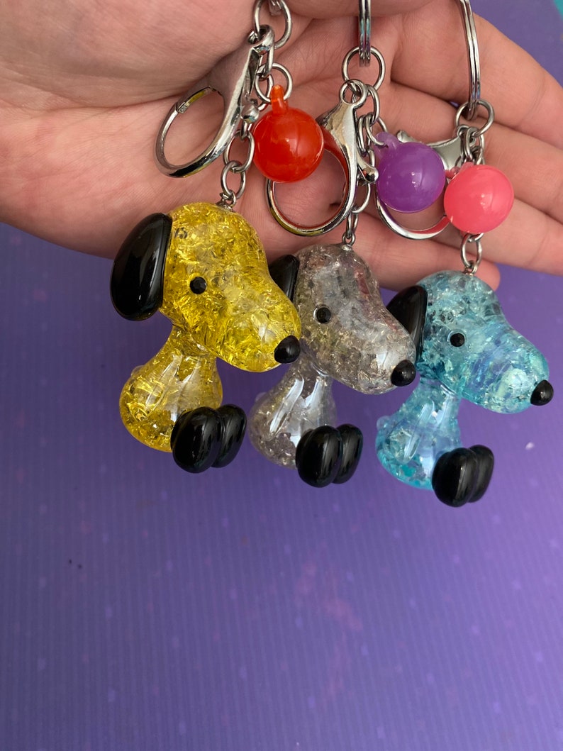 May include: Three keychains featuring a cartoon dog design. The dogs are made of clear glass with glitter embedded inside. The dogs are yellow, gray, and blue. Each keychain has a silver ring and a small, colorful ball charm.