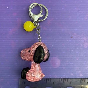 May include: Pink and black glass charm of a cartoon dog with a yellow ball keychain attached. The charm is about 1 inch tall.