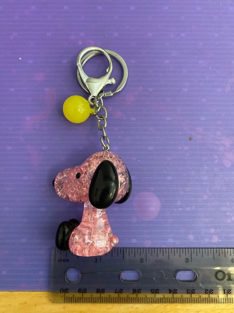 May include: Pink and black glass charm of a cartoon dog with a yellow ball keychain attached. The charm is about 1 inch tall.