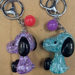May include: Two keychains featuring a cartoon dog with a sparkly, translucent body. One keychain is purple with a red bead, and the other is blue with a purple bead.