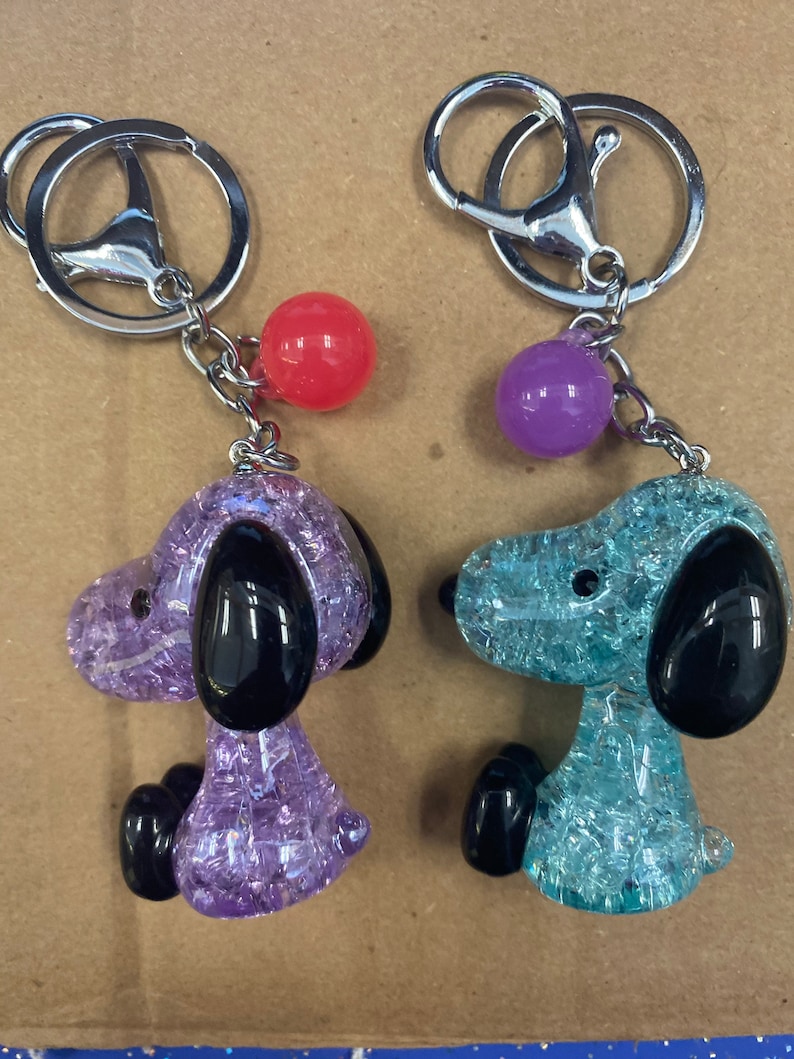 May include: Two keychains featuring a cartoon dog with a sparkly, translucent body. One keychain is purple with a red bead, and the other is blue with a purple bead.