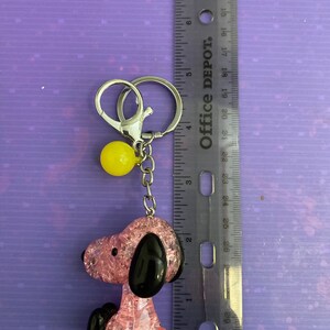 May include: Pink and black glitter charm of a cartoon dog with a yellow ball charm attached to a silver key chain.