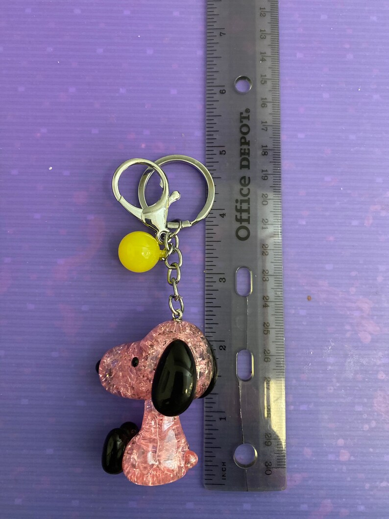 May include: Pink and black glitter charm of a cartoon dog with a yellow ball charm attached to a silver key chain.