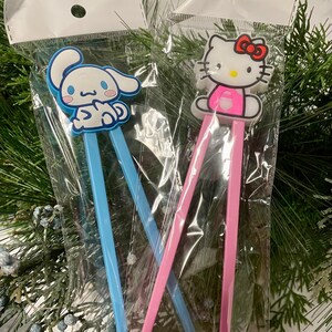 May include: Two pairs of chopsticks with cartoon character toppers. One pair has a blue handle and a white bunny character topper. The other pair has a pink handle and a pink Hello Kitty character topper.