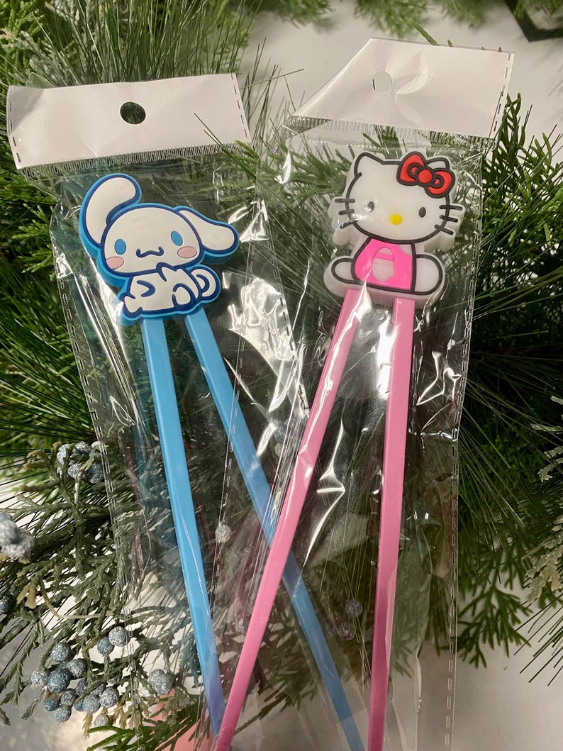 May include: Two pairs of chopsticks with cartoon character toppers. One pair has a blue handle and a white bunny character topper. The other pair has a pink handle and a pink Hello Kitty character topper.