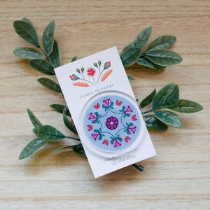 Peut inclure: Un pin's circulaire sur fond bleu clair avec un motif floral symétrique en forme de mandala dans des tons de rose, violet et vert. Le pin's est fixé sur une carte de présentation blanche portant le texte FLORAL FEATHERS et www.floralfeathers.ca.
