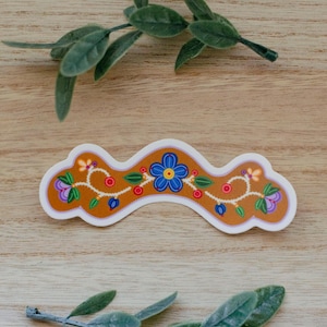 Peut inclure: Un autocollant ondulé découpé à la forme, présentant un fond jaune moutarde avec une fleur bleue centrale, entourée de vignes blanches entrelacées, de petites baies rouges, de feuilles vertes et d'accents floraux violets, le tout bordé de blanc.