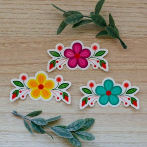 Peut inclure: Trois autocollants découpés présentant des motifs floraux en magenta, jaune et sarcelle. Chaque autocollant affiche une fleur centrale à cinq pétales avec des feuilles vertes, des baies rouges et des lignes pointillées sur fond blanc.
