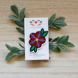 Peut inclure: Une broche florale représentant une fleur rouge, bleue et jaune avec des feuilles vertes sur une carte de présentation blanche. La carte affiche le texte Floral Feathers et www.floralfeathers.ca. Le motif inclut des points et des pétales.