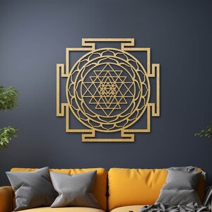 Op de afbeelding: Een gouden houten wanddecoratie met een geometrisch ontwerp met ingewikkelde details. De decoratie hangt aan een donkerblauwe muur in een woonkamer met een gele bank en planten.
