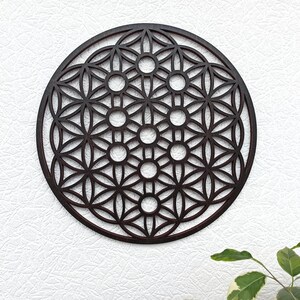 May include: A dark brown wooden wall hanging in the shape of a circle with a geometric pattern of overlapping circles and flower shapes. The pattern is known as the Flower of Life.