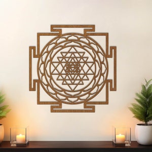 Op de afbeelding: Een bruine houten wanddecoratie met een geometrisch ontwerp, met een Sri Yantra, een heilig symbool in het hindoe&iuml;sme. De wanddecoratie is te zien op een witte muur met twee kleine potplanten en kaarsen op een houten plank.