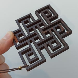 May include: A dark brown wooden pendant with an intricate, geometric knot design. The pendant is held by a hand and has a brown cord attached. The design is a complex series of interlocking squares and lines.