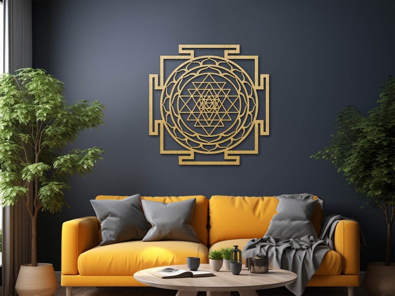 Op de afbeelding: Een gouden houten wanddecoratie met een geometrisch ontwerp met ingewikkelde details. De decoratie hangt aan een donkerblauwe muur in een woonkamer met een gele bank en planten.