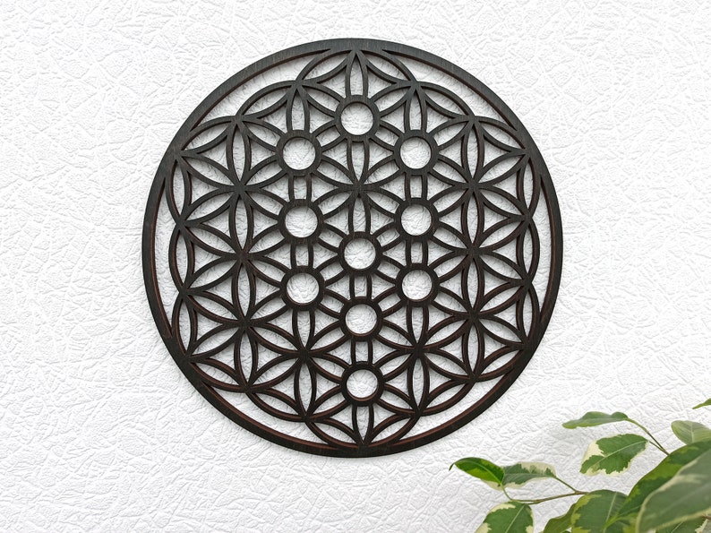 May include: A dark brown wooden wall hanging in the shape of a circle with a geometric pattern of overlapping circles and flower shapes. The pattern is known as the Flower of Life.